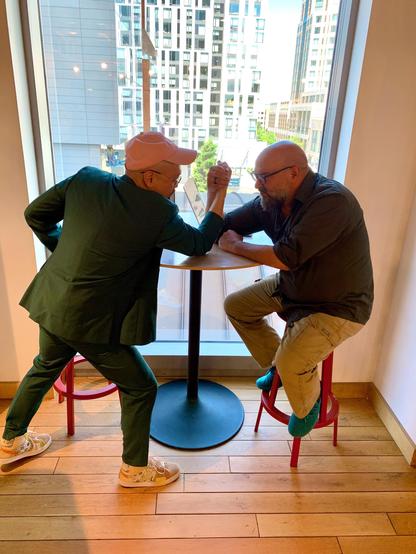 Ken and Jeff engage in an arm wrestling match.  Ken is standing. Jeff remains seated on a bar stool