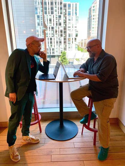 Ken and Jeff are on opposite sides of a bar table. Both have their laptops open. Ken stands on the left with his elbow on the table and chin in his hand. Jeff sits on a bar stool and smiles as he types. The window behind them reveals high-rises and sunshine in the Oakland cityscape