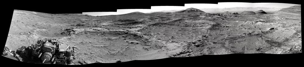 A mosaic of eight black and white images capturing a Martian landscape, with a section of the rover in the foreground to the left.