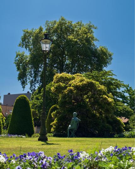 A beautifully maintained garden with a vintage street lamp in the foreground and various manicured plants. A sculptural figure can be seen sitting in a chair under the shade of a tree. The scene features carefully trimmed topiaries, flowering plants in purple and white at the bottom of the frame, and a clear blue sky above. The composition creates a serene park setting with classical garden elements.