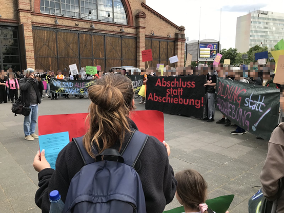 Menschen stehen im Kreis zur Auftaktkundgebung vor dem Bockenheimer Depot in Frankfurt am Main. Die meisten sind Schüler*innen. Sie haben Transpis dabei:
- Abschluss statt Abschiebung!
- Bildung statt Abschiebung!
- Wir sind alle mit Angad & Gunit!
Viele Menschen halten auch Schilder hoch.