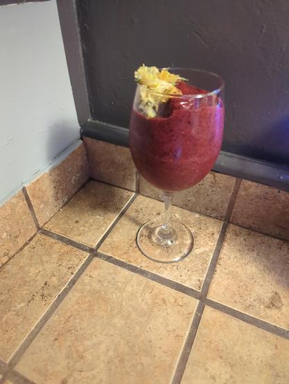 A smoothie with honeysuckles on top inside a wine glass sitting on a brown tile counter. Tile comes up the wall a bit, then turns to a grey wall as you look up. Wineglass shadow casted on a lighter grey wall