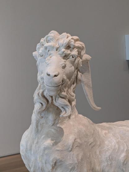A marble sculpture of a goat. It has wild eyes, a closed mouth grin and curls for days.