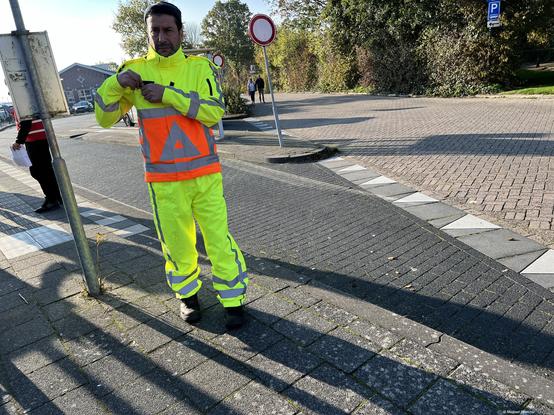Photo by Fenichel

A bright "human warning sigh" - a man in florescent.yellow-green jupsuit with his midsection featuring a big square bright-orange sign with the universal (triangle) warning sign - is standing on asphalt on what appears to be a crosswalk but is banda of shadow from poles. He is on the left half of the frame while the right side is another pattern-rich street of cobblestone, punctuated by white edgings.