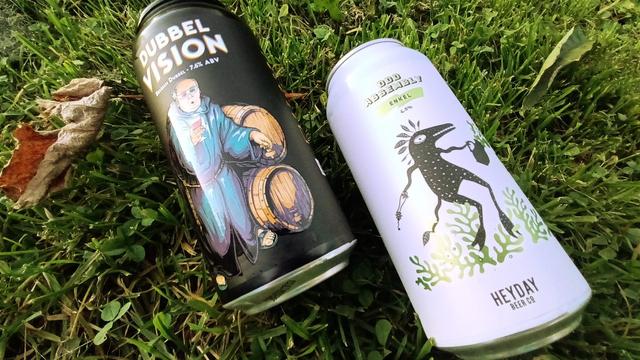 Two 440ml beer cans lying in the grass, label side up. 

On the left: Double Vision Dubbel Vision Belgian Dubbel - black label with a cartoon-ish monk in front of barrels. Text is wobbly, like fuzzy vision, seeing double.

On the right: Heyday Enkel Odd Assembly Belgian Ale - white label with black text and ditto cartoon figure, like a stick man or blob. Green accents in the back, grassy.