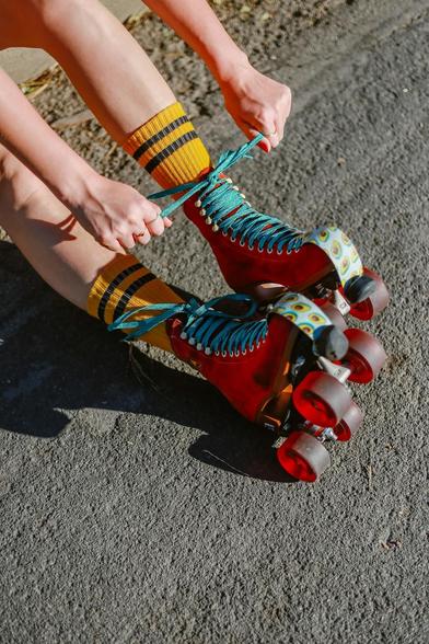 Man sieht rote Retro-Rollschuhe auf Asphalt mit blauen Schnürsenkeln. Zwei Hände binden die Schnürsenkel gerade.