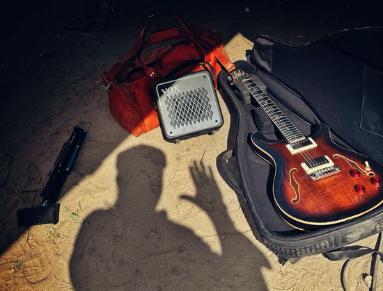 Ein Foto meines Equipments (Gitarre, Verstärker und Tasche) auf sandigem Boden mit Schatten meinerselbst, die Hand winkend erhoben.