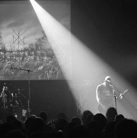 Een zwart-witfoto van een liveoptreden van een metalband. De zanger/gitarist staat rechts op het podium onder een felle spot, terwijl op de achtergrond een projectie te zien is van een veld met gras en bomen en een mystiek symbool. Links zit de drummer achter zijn drumstel. Het publiek is zichtbaar op de voorgrond.