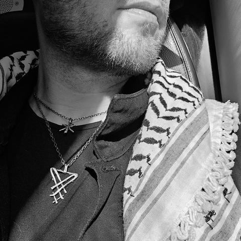 Zwart-wit close-up van de onderkant van een gezicht van een persoon met een korte baard. De persoon draagt twee kettingen met hangers: één met een sigil van Lucifer en één met een bindrune die 'Liefde' symboliseert. Over de schouder hangt een keffiyeh met het traditionele zwart-wit patroon. De persoon kijkt weg van de camera en zit mogelijk in een trein.