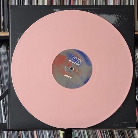 Auf einem schwarzen Plattenspieler liegt eine rosafarbene Schallplatte mit einem Etikett, auf dem "Ditz" und "Side B" steht. Im Hintergrund ist eine Sammlung von anderen Schallplatten zu sehen.