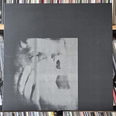 Ein teilweise verdecktes Schwarz-Weiß-Albumcover mit einem stilisierten Bild des Gesichts einer Person mit einem Ausdruck von Schock oder Überraschung. Der Hintergrund ist überwiegend schwarz, und das Album wird vor einem Hintergrund aus Schallplatten gezeigt.