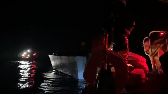 Dunkle Nacht. Von einem RHIB aus ist voraus ein treibendes nicht sehr großes Holzboot zu sehen, auf dem Menschen dicht zusammen gedrängt sind. Auf der anderen Seite des Bootes ein weiteres RHIB.