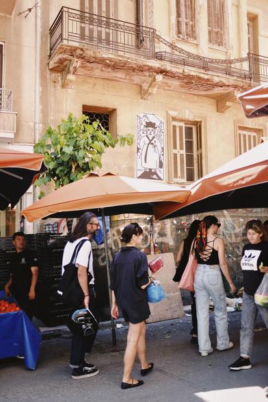 People walking through an outdoor farmers market with bright orange umbrellas, set in front of an old building with peeling paint and a black-and-white street art poster of a tiger strolling down the market scene.