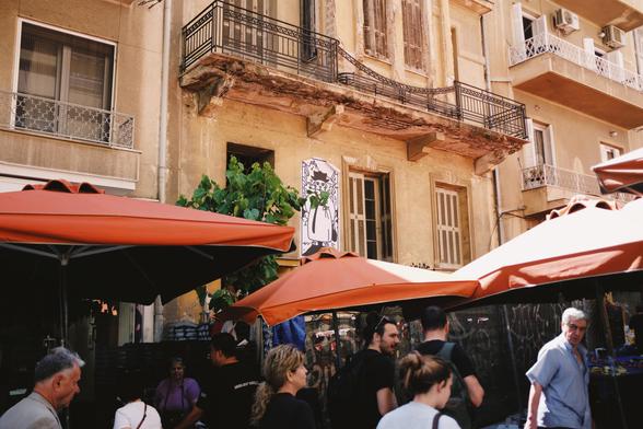 People walking through an outdoor farmers market with bright orange umbrellas, set in front of an old building with peeling paint and a black-and-white street art poster of a tiger strolling down the market scene.