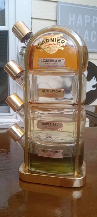 A four tiered bottle in a brass stand. The bottles are stacked upon one another and labeled, Liqueur D'or, Creme de Cacao, Triple Sec and Creme de Menthe. Alcohol is in all the bottles except the creme de cocao. The top bottle has a label  that says. "Garnier Liqueur Enchien Paris, France." The bottles are held together with two brass arcs that travel up each side, around the top and back down again. The top is rounded and the bottles are mounted on a brass and plastic base. 