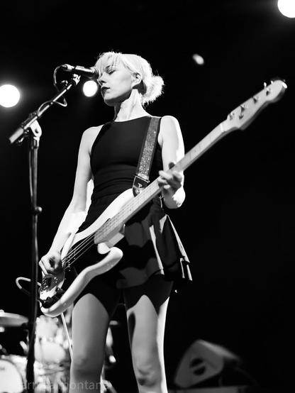 A female musician is performing on stage, playing a white bass guitar. She wears a black dress and stands in front of a microphone, with focused expression and bright stage lights in the background. The image is in black and white.