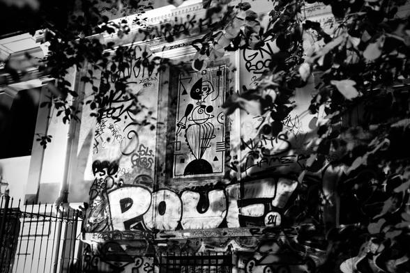 Black and white image of a building facade covered in graffiti, showcasing a geometric tiger paste-up created as an homage to Oskar Schlemmer’s Triadic Ballet, partially hidden behind tree branches.