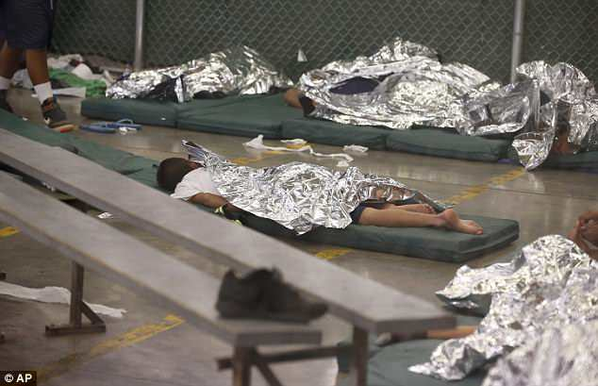 A color photo of children sleeping under mylar survival blankets on thin mattresses, on the floor of a detention facility. The are appears to be enclosed by chain-link fencing.