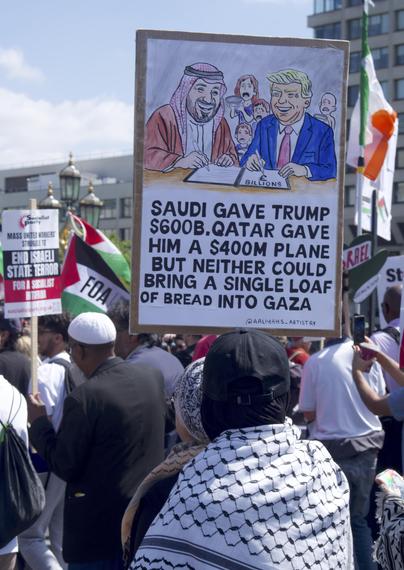 woman carrying sign "Saudi gave Trump $600B.QATAR GAVE HIM A $400M PLANE BUT NEITHER COULD BRING A SINGLE LOAF OF BREAD INTO GAZA "

