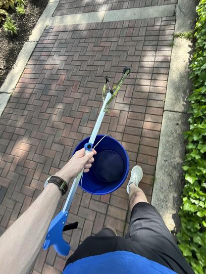 A photograph taken of a person from just above their waist pointing toward the ground. They are holding a bucket and grabber tool. A thin strip of green plants lines the right side, and a brick path fills the majority of the frame. 