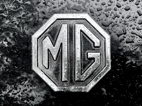 Photograph of an MG car logo consisting of a capital MG in an octagon. The logo is on a black car and covered by raindrops.