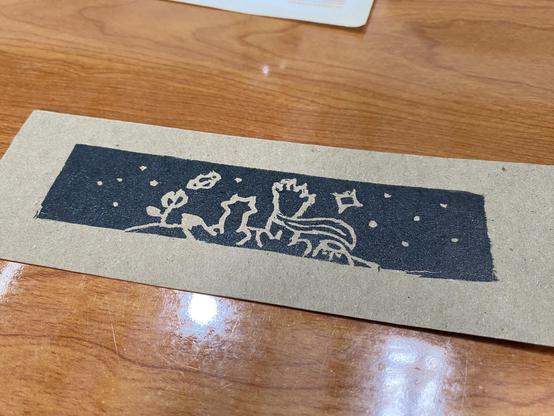 Fragmento de papel café con un estampado negro, rectangular, en donde destaca la silueta de El Principito, el zorro y una rosa viendo a las estrellas.