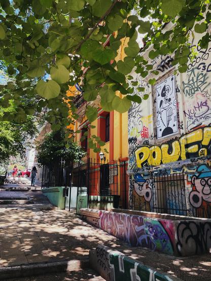 Vibrant street scene in Athens featuring a graffiti-covered neoclassical building with a black-and-white geometric tiger paste-up in a window frame, inspired by Schlemmer’s Triadic Ballet, partly hidden by lush tree branches.