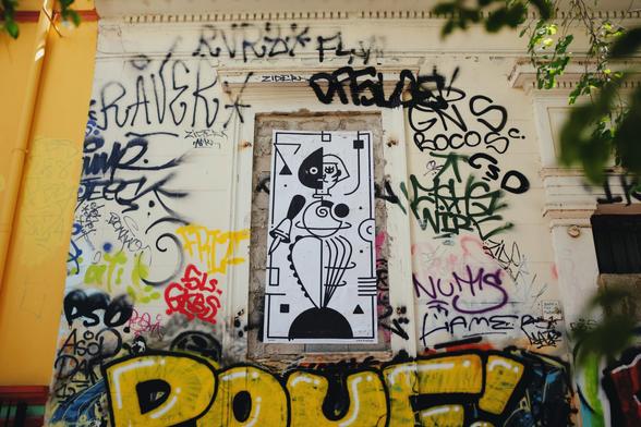 Close-up of a graffiti-covered neoclassical building with a black-and-white geometric tiger paste-up in a window frame, inspired by Schlemmer’s Triadic Ballet, partly hidden by lush tree branches.