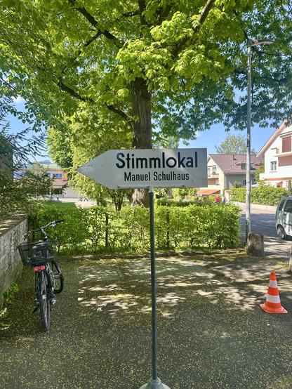 Ein Info-Ständer mit Aufschrift: «Stimmlokal, Manuel Schulhaus» steht auf dem Platz. Dahinter ein schön grüner Baum und ein abgestelltes Fahrrad.