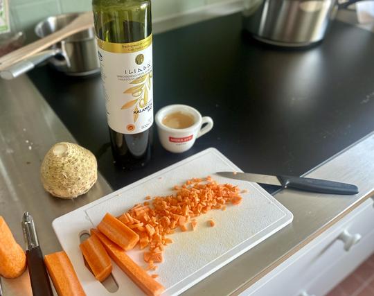 Küche/Anrichte. Ein weisses Kubststoffbrett mit Karotten in verschiedenen Grad der Zerkleinerung. Links eine Knollensellerie. Neben der Flasche Olivenöl mit Aufschrift: Kalamata PDO steht eine Tasse Espresso mit roter Aufschrift Moka Efti. Rüstmesser liegen links und rechts des Bretts.