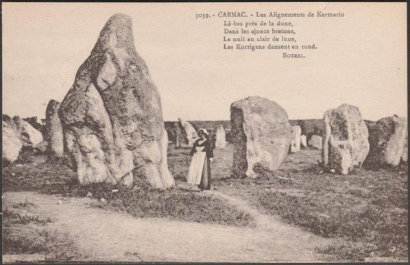 A vintage printed postcard of the Alignements de Kermario, a series of rows of standing stones or menhirs near Carnac in Brittany. The rows of stones lead off into the distance and a much larger menhir, the Géant de Kermario, is at the front left. A young woman in traditional Breton costume stands beside it on a rough path. Four lines from Botrel's La Bonne Chanson is at the top right, and reads: "Là -bas,  près  de  la  dune, Dans  les  ajoncs  bretons, La  nuit  au  clair  de  lune, Les  Korrigans  dansent  en  rond.", roughly translated as "Over there, near the dune, In the Breton gorse, At night in the moonlight, The Korrigans dance in a circle."