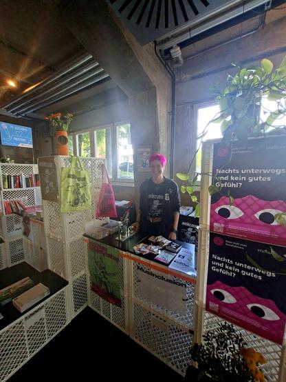 Eine Frau mit kurzen pinken Haaren steht an einem Stand des About Pop Festivals. Der Stand ist aus weißen Bäckerei-Kisten gebaut und mit Werbematerial, Plakaten und bunten Taschen "dekoriert". Oben stehen links und rechts Blumen auf dem Aufbau. Im Hintergund sind Fenster zu sehen