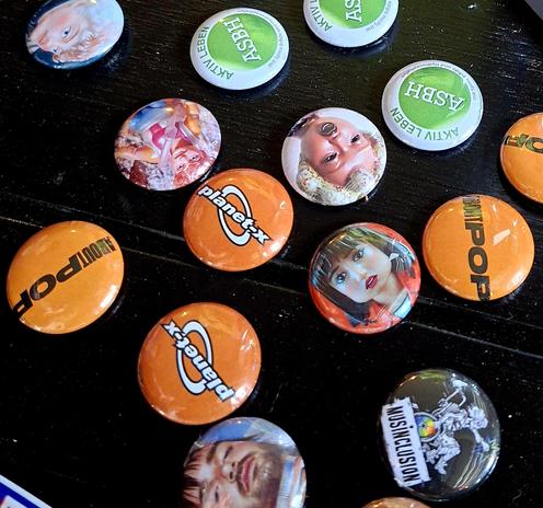 Bunte Buttons liegen auf einem schwarzen Tisch. Es sind Gesichter zu sehen, aber auch Logos von MusInclusion, About Pop und dem Jugendhaus planet-x