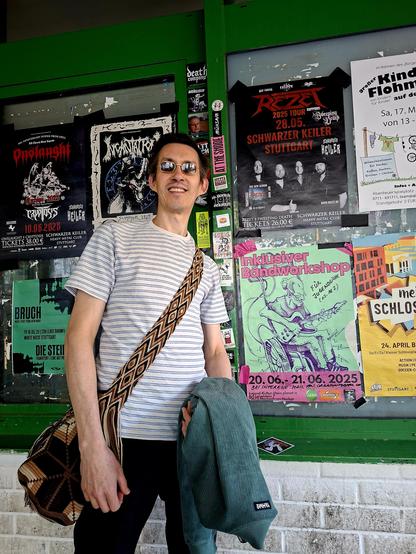 Ein großer Mann mit dunklen Haaren und Sonnenbrille steht vor einer plakatierten Wand. Rechts neben ihm das Plakat des Inklusiven Bandworkshops