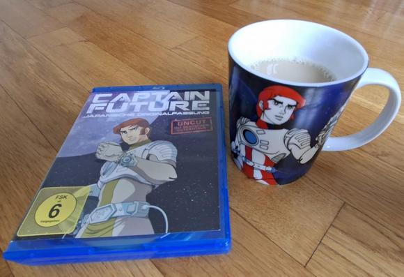 Das Foto zeigt eine gefüllte Captain-Future-Kaffeetasse sowie die BluRay-Ausgabe der japanischen Fassung (ungeschnitten) der TV-Serie, beides plaziert auf einem hellen Parkettboden...
