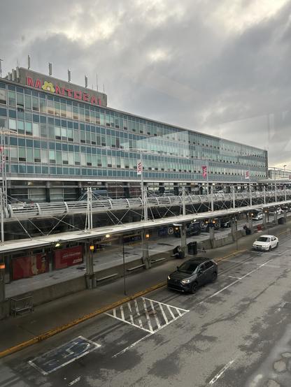 A view of the Montréal-Pierre Elliott Trudeau International Airport terminal, showcasing a modern glass building under a cloudy sky. The image includes a busy pickup area with parked vehicles and a few people waiting.