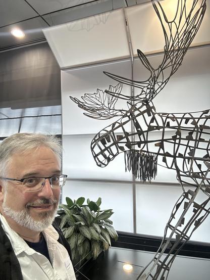 An interior scene featuring a smiling man with glasses next to a large wireframe sculpture of a moose. There is also a green plant and soft lighting present in the background.