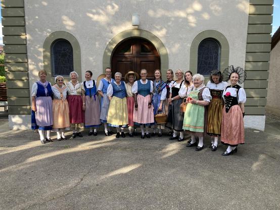 Fifteen women in assorted Swiss cantonal costumes stand in a line in front of a Church. They are smiling and basically happy & relieved to have survived their concert despite their musical director & professional singer soloist having been unable to do more than mouth the words!