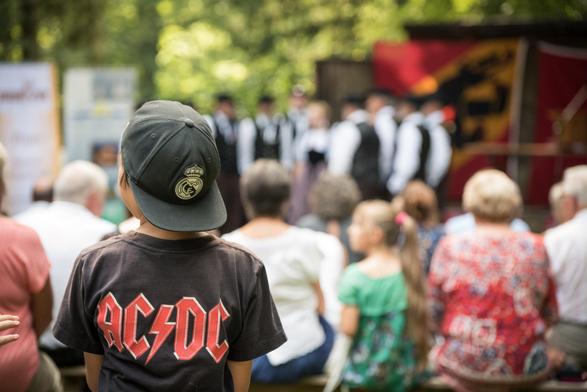 Unscharfe Fotografie eines Dorffestes mit Jodlerchor, Bernerfahne und Publikum; im Vordergrund scharf ein Kind von hinten mit einem AC/DC-T-Shirt.