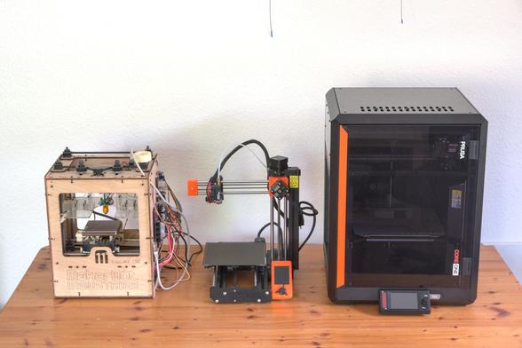 3 3D-Drucker auf einem Tisch. Der Linke ist aus gelaserten Sperrholz und Kistenförmig. Der Mittlere ein offener "Bedslinger", der Rechte eine grosse Kiste aus  gestanzten Blech (20kg).