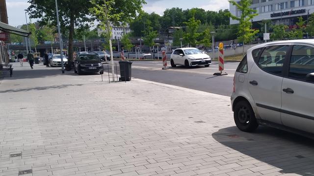 Foto zeigt neu gestalteten Gehweg und straßenbegleitende Parkplätze an der Friedrichstraße in Düsseldorf. Die Fläche ist übergangslos einheitlich mit hellgrauen rechteckigen Pflastersteinen ausgestattet. Die Baumscheiben eines neuen Baums ist ein winziges Loch im Pflaster, dazu markierte, besetzte Parkplätze und ein paar Fahrradständer. Im Hintergrund ein Einkaufszentrum.