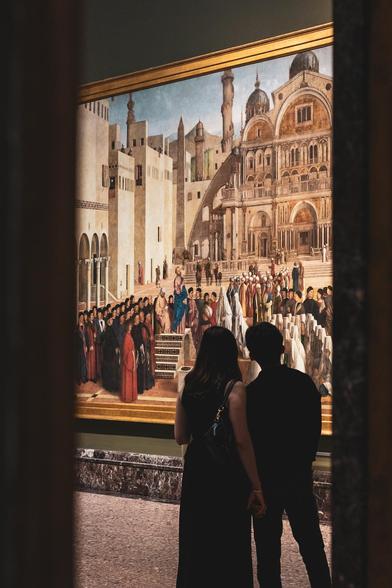 Fotografía vertical donde se ve una pareja observando la Predicación de San Marcos en Alejandría de Giovanni y Gentile Bellini en la Pinacoteca de Brera (Milán).