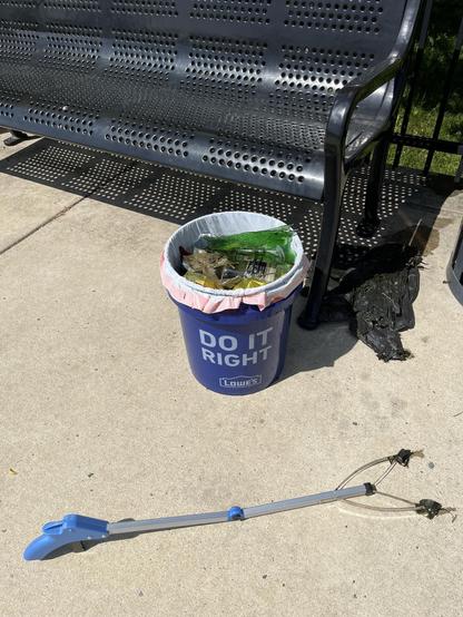A five gallon bucket in front of a bench. The bucket is filled with trash recovered from and around a pond. A small trash grabber lays in front of it.