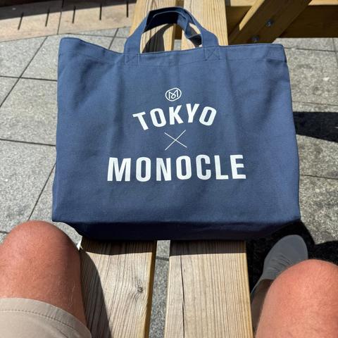 Close up of a navy blue canvas tote bag, which reads “Tokyo x Monocle” in white text. It’s resting on a bench in front of me and my knees are just visible poking out of my shorts - sorry! lol 