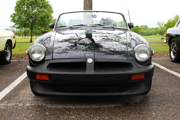 Photo of the front end of a black 1976 MG MGB on a rainy day in the parking lot of a park.