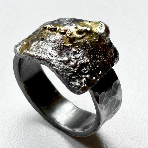 A unique, textured ring featuring a rough, metallic design with a small gold chain accent on top. The band has a hammered finish, adding an artisanal touch.
