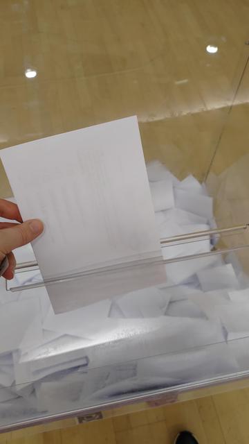Zdjęcie przedstawia złożoną kartę do głosowania, częściowo wsuniętą  do przezroczystej skrzynki wyborczej. Dno skrzynki pokryte jest białymi złożonymi kartkami.
