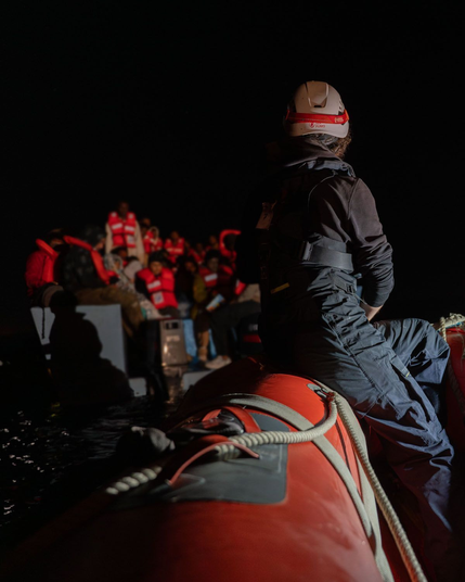 Eine Rettungskraft mit Helm und Schutzausrüstung sitzt auf dem Rand eines roten Schlauchbootes und blickt auf ein überfülltes Boot mit Menschen, die orangefarbene Rettungswesten tragen, bei Nacht auf dem Wasser.