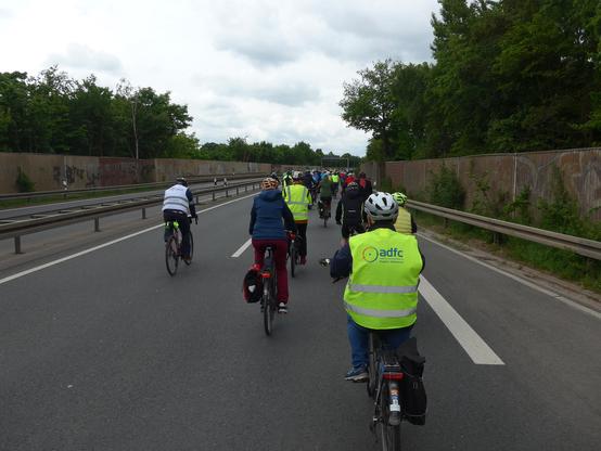 Viele Fahrradfahrer von hinten auf einer zweispurigen Bundesstraße, im Vordergrund ein Radfahrer mit Warnweste mit Aufdruck ADFC Region Hannover, links ein Ordner mit weißer Warnweste