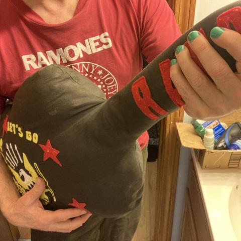 a guitar-shaped pillow decorated with the Ramones insignia being "played by a man in a red Ramones shirt with teal-colored finger nails